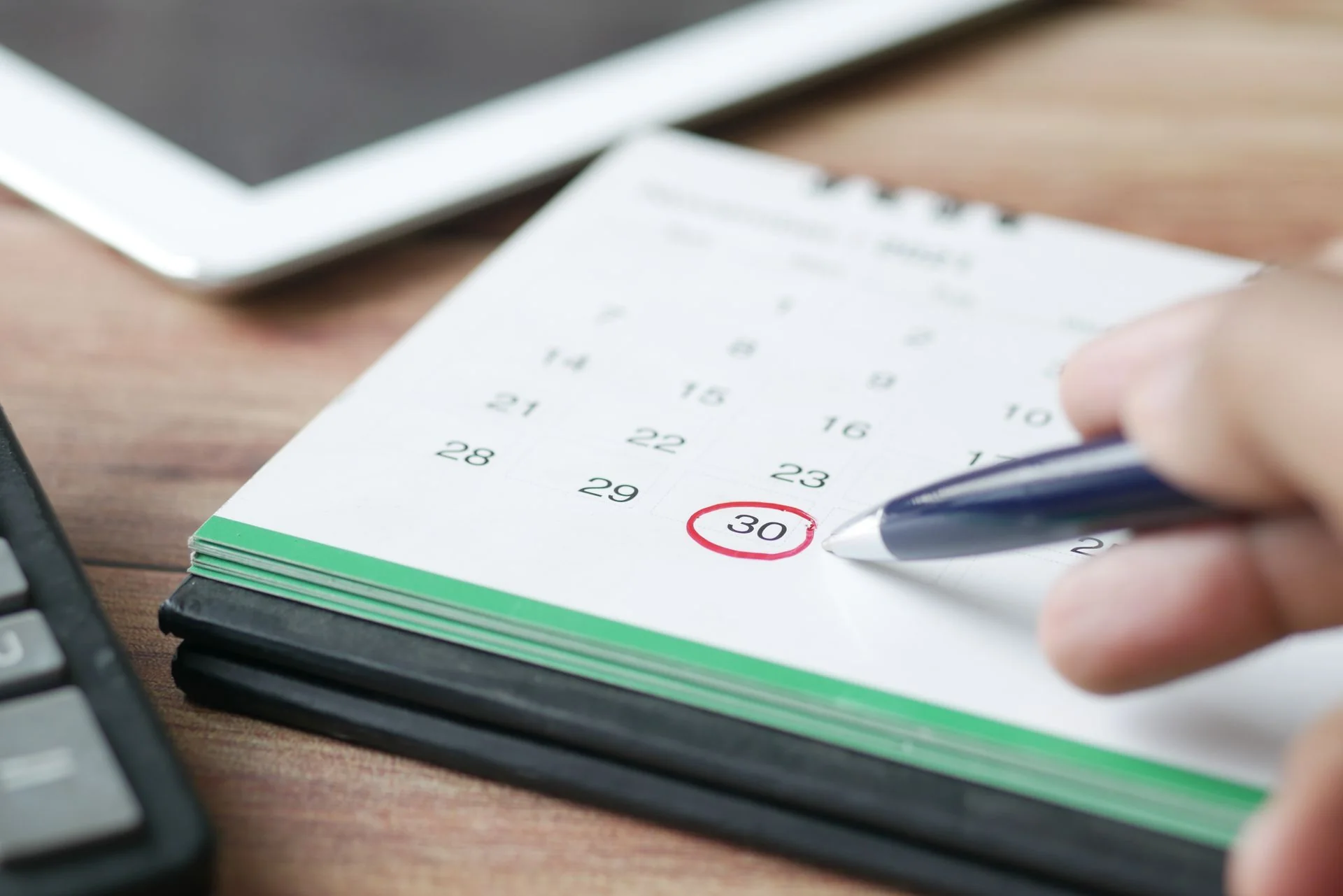 A hand circles a date on a paper calendar beside a tablet and keyboard.