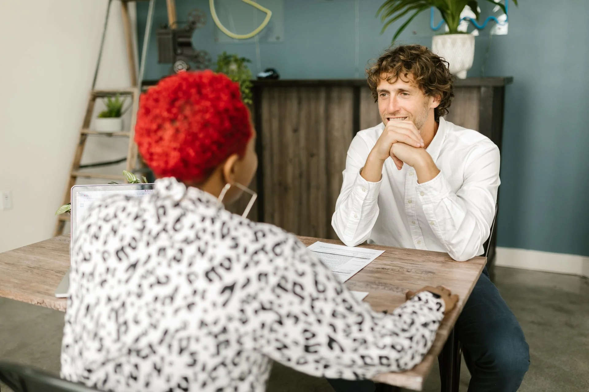 Two people sitting across a wooden table in conversation, one with their hands clasped.