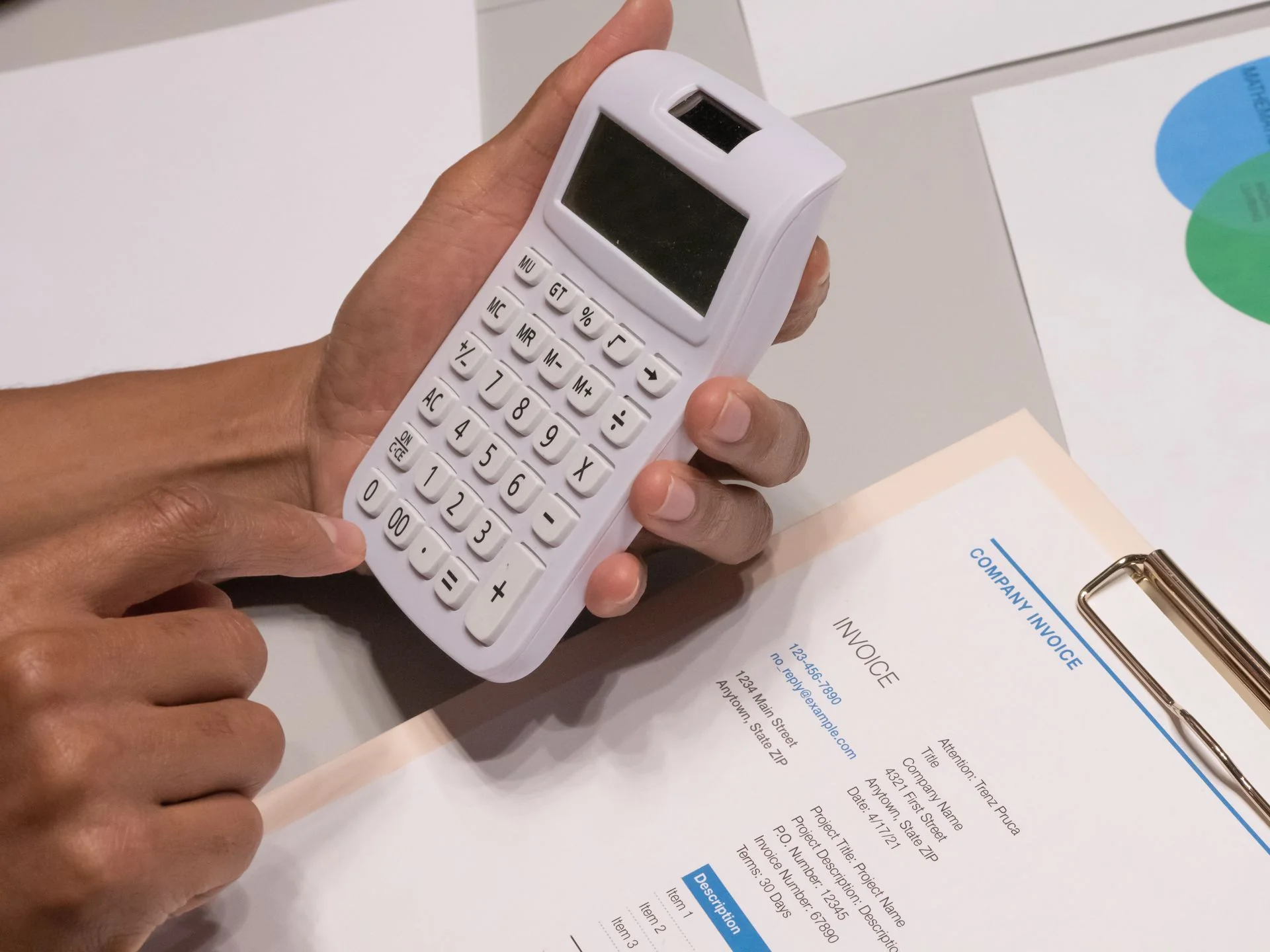 Hands using a calculator beside an invoice on a clipboard.