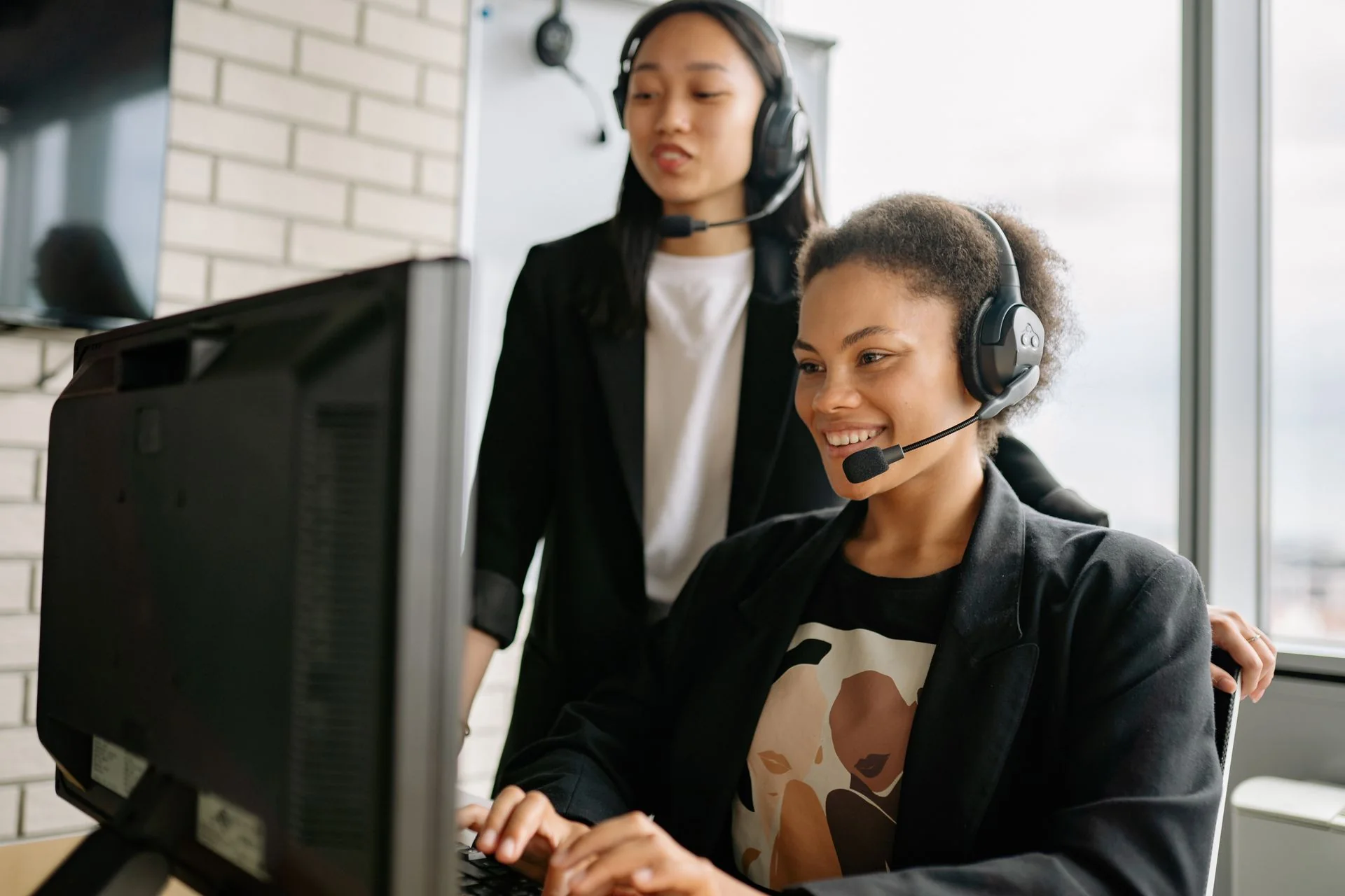 Two people wearing microphone headsets work together at a computer.