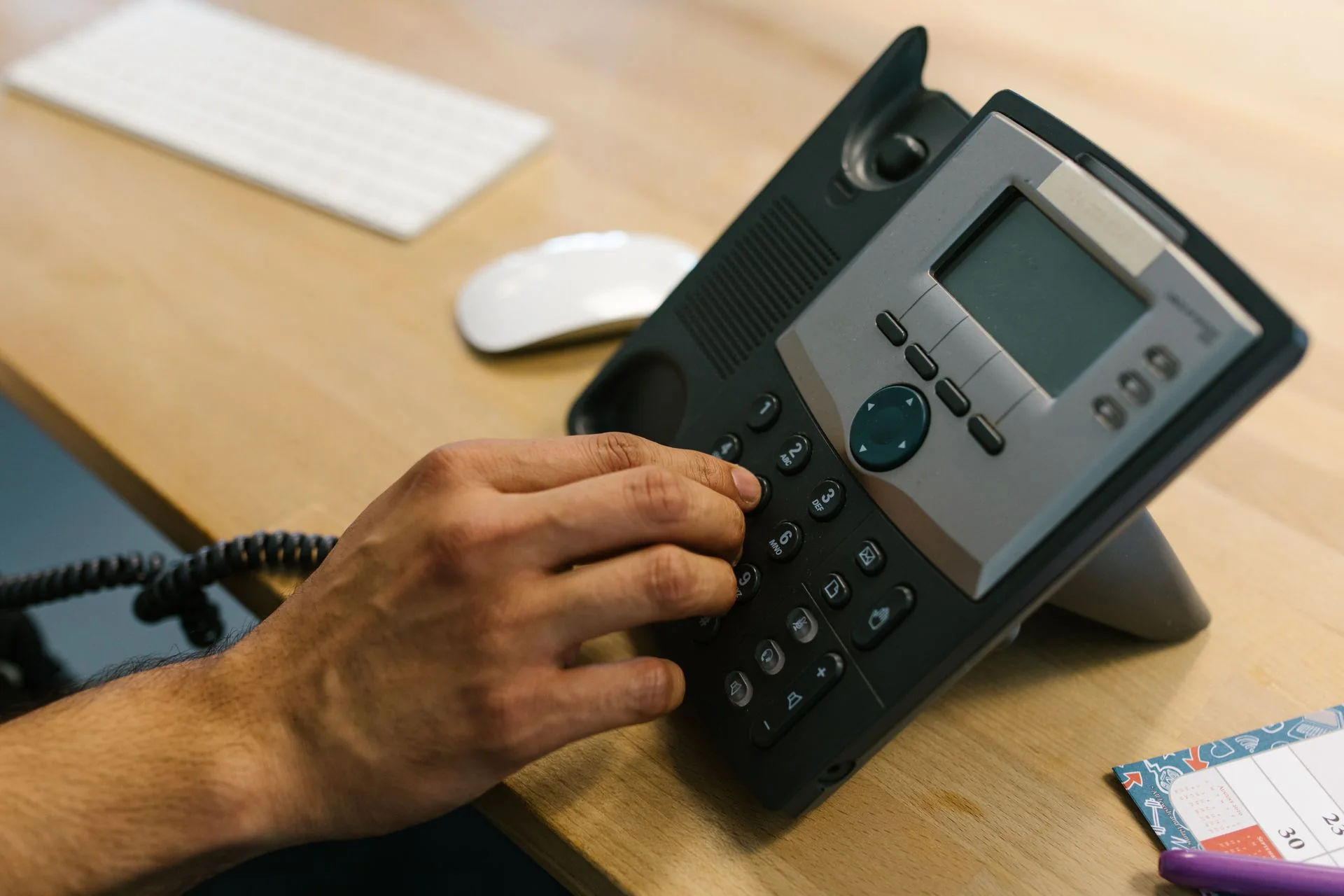 A hand pressing the keypad of a black desk telephone beside a keyboard and mouse.