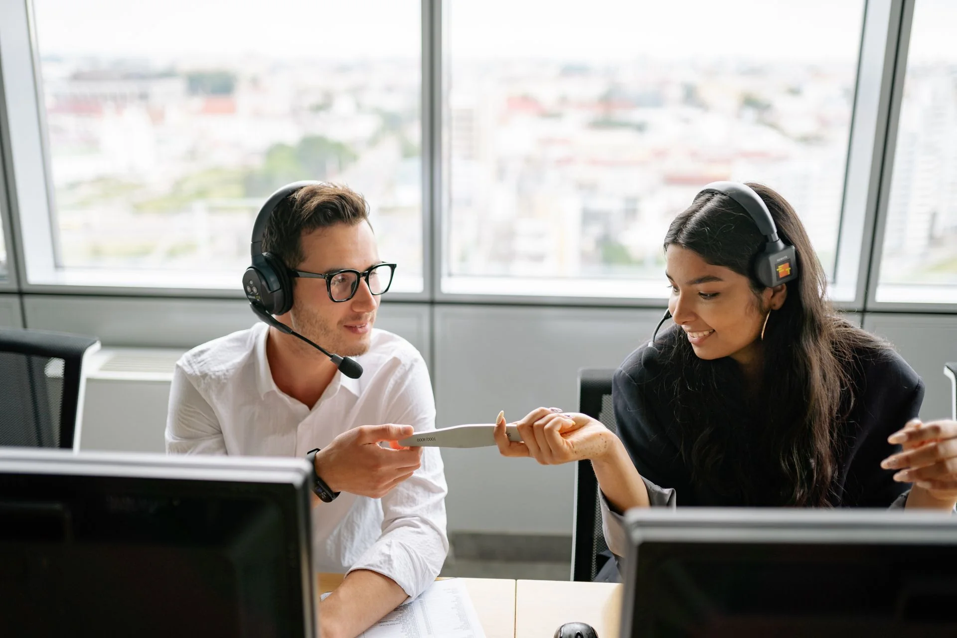 Two headset-wearing office workers discussing a paper document between computer monitors.