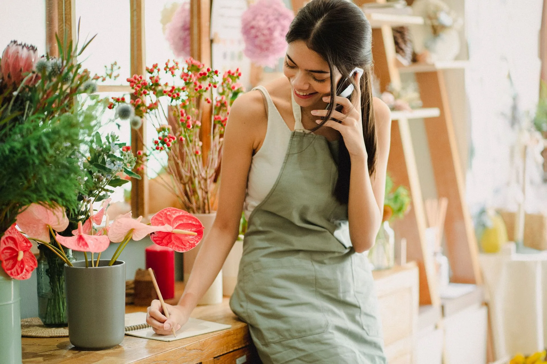 Florist speaking on a mobile phone and writing notes at a flower-shop counter.