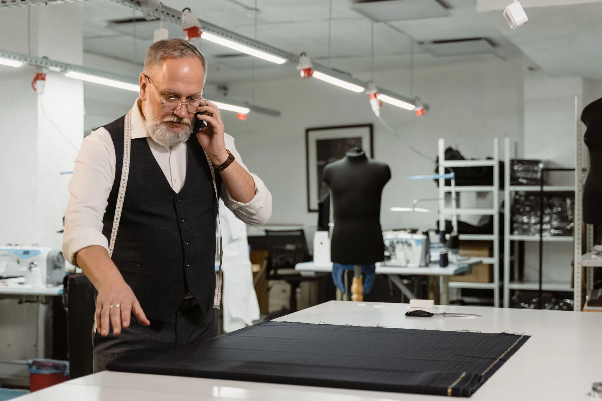 Tailor examining fabric on a workshop table while speaking on a mobile phone.