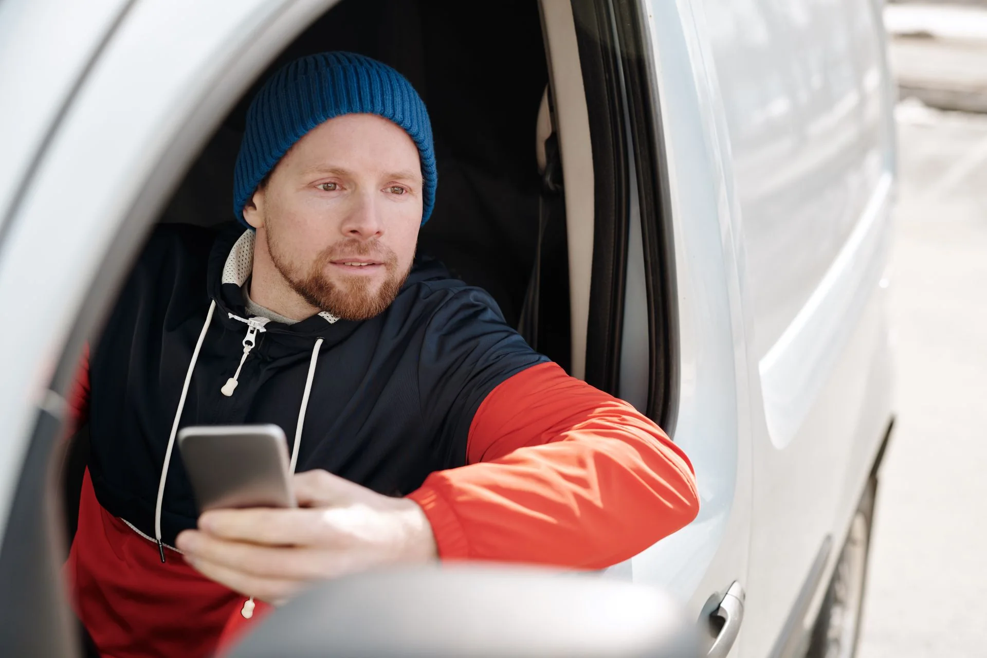 A man in a beanie sitting in the open door of a white work van, looking at his phone.