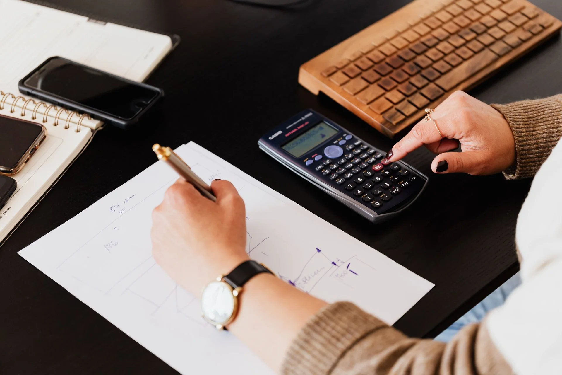 Hands using a calculator and writing figures beside a keyboard and notebook.